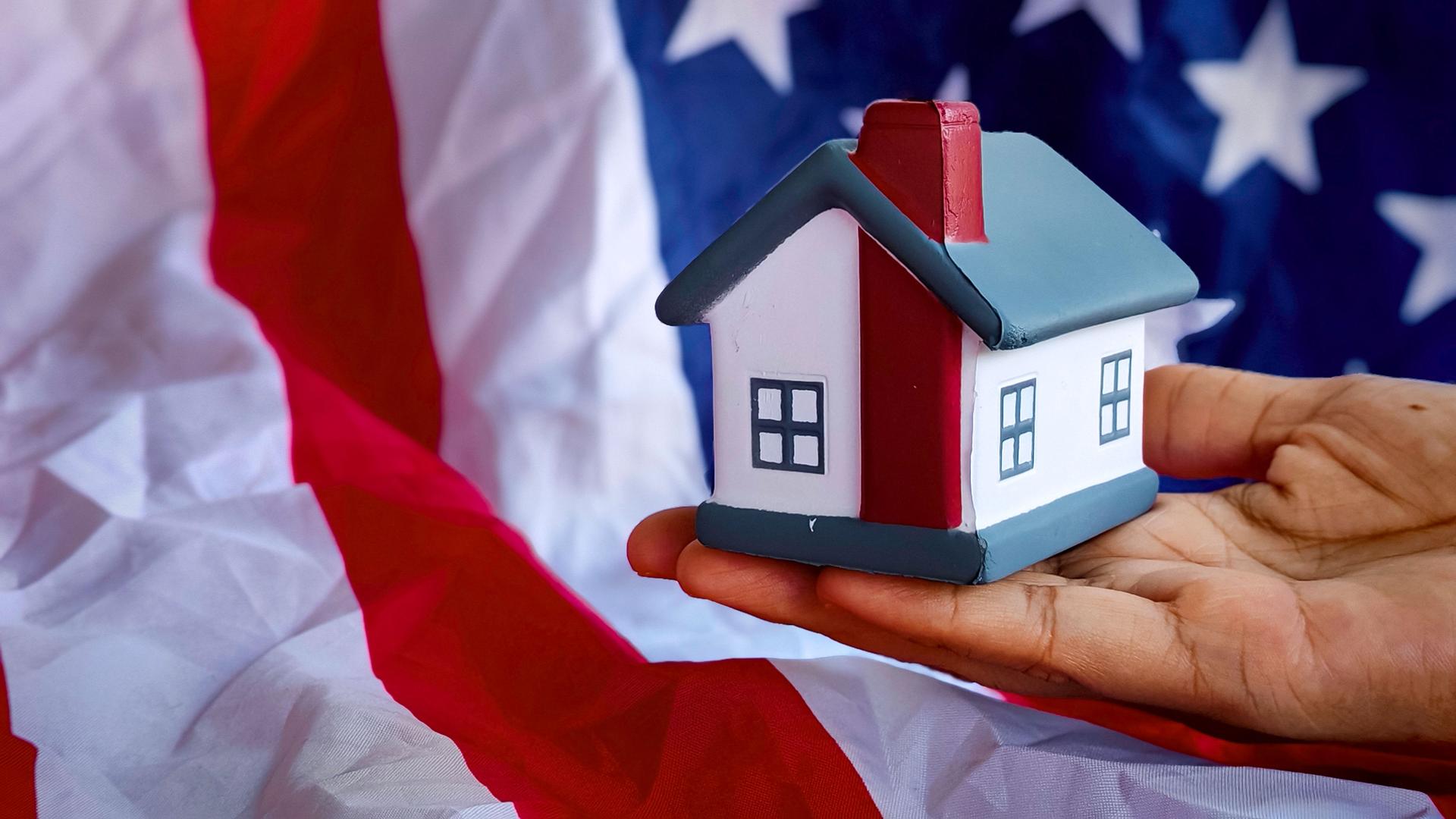 A hand holding model house with background of usa flag.